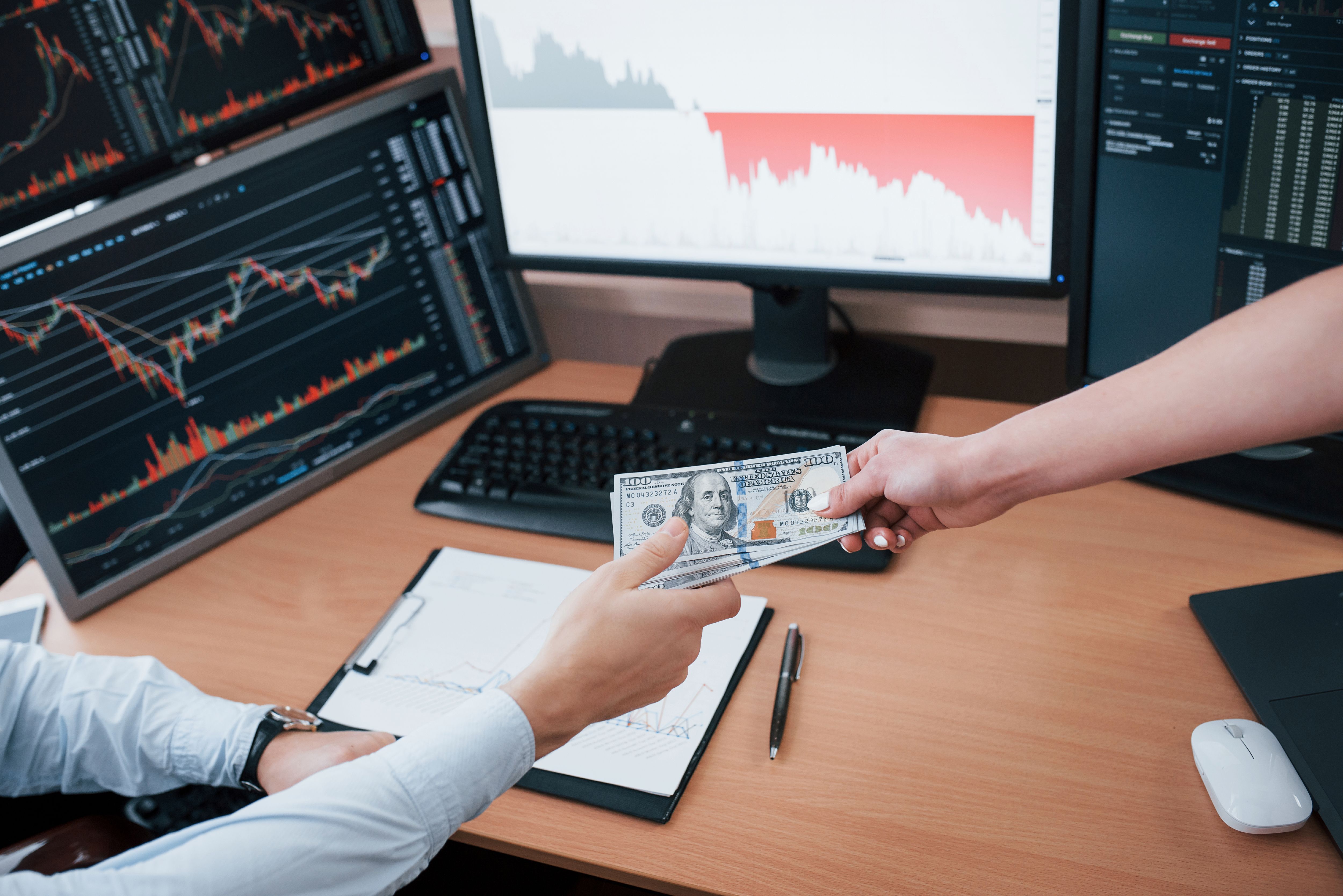 Hands of people passing dollars to each other against the background of monitors with graphs