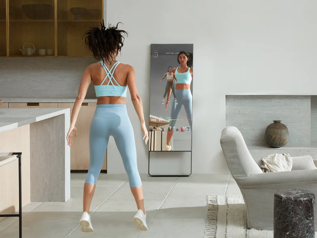 Young woman is jumping in her apartment in front of The Mirror device, which reflects her, displaying her instructor and other participants