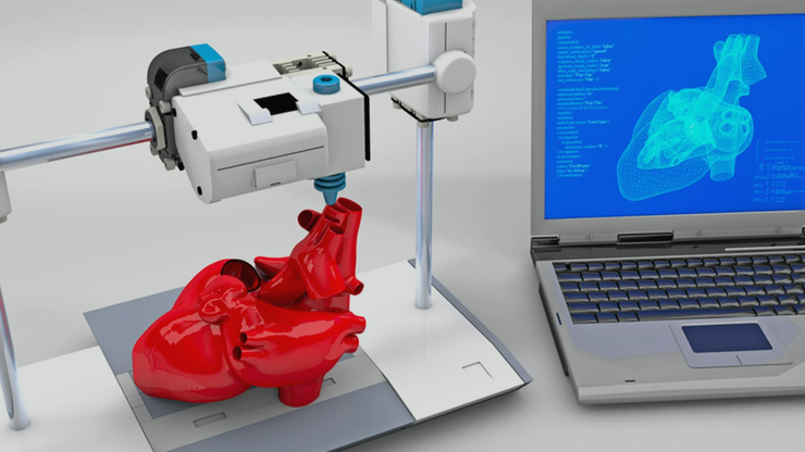 3D printer printing a red human heart and a laptop standing nearby with 3D model of this heart on the screen